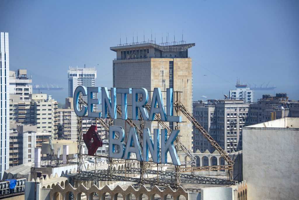 A large “Central Bank” sign mounted on top of a building, overlooking a dense urban cityscape with high-rise buildings and the sea in the background.