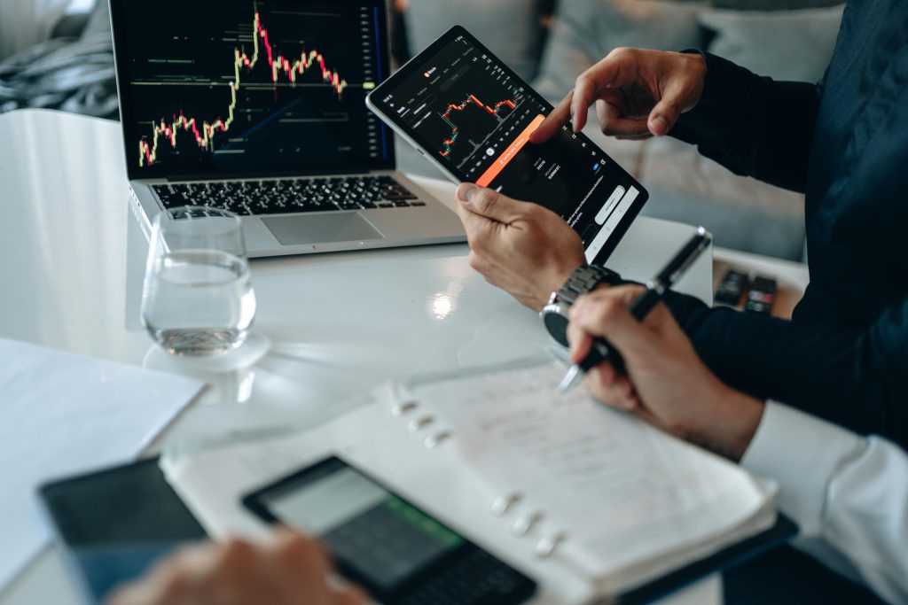 Two people analyzing financial charts on a laptop and smartphone, reviewing market data with notes and a calculator on the table.