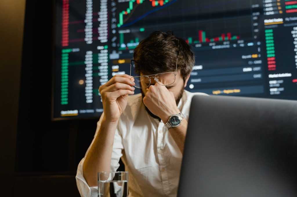 Trader sitting at a laptop in front of large trading screens with market charts, rubbing his eyes and holding his glasses, appearing stressed or fatigued.