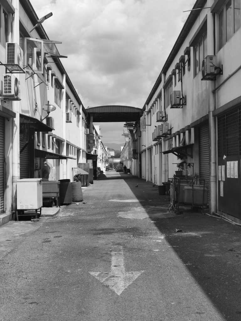 Black-and-white photo of a narrow alley between industrial buildings with air conditioning units, bins, and a covered walkway in the distance.